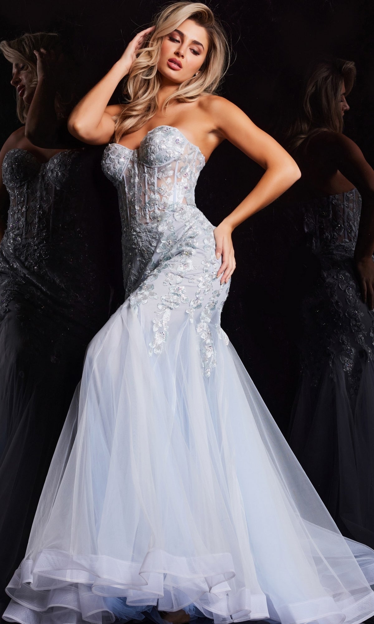 A woman poses against a dark reflective background wearing the Jovani Strapless Long Light Blue Prom Dress 22924—a modern, fitted mermaid gown featuring a sheer corset bodice, floral embroidery, and a flowing tulle skirt.
